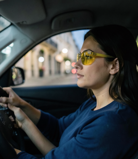 Femme au volant portant des lunettes de conduite à verres jaunes dans une ville animée.
